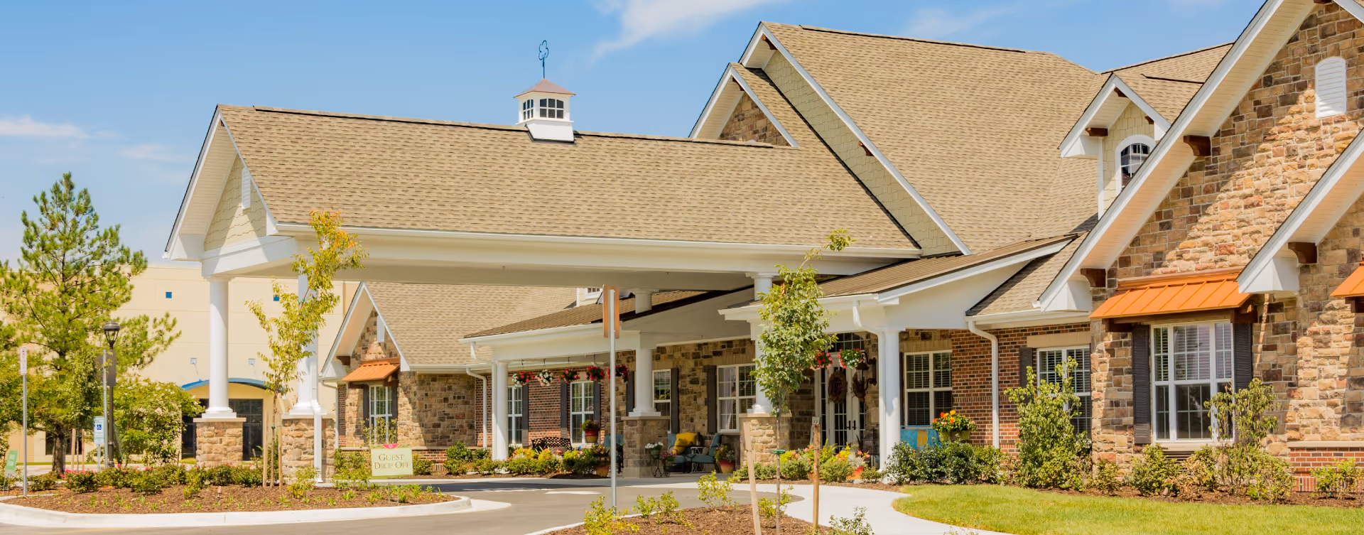 Exterior view of Bickford of Suffolk senior living facility showing a covered entrance with white columns, stone and brick facade, landscaped greenery, and a clear blue sky.