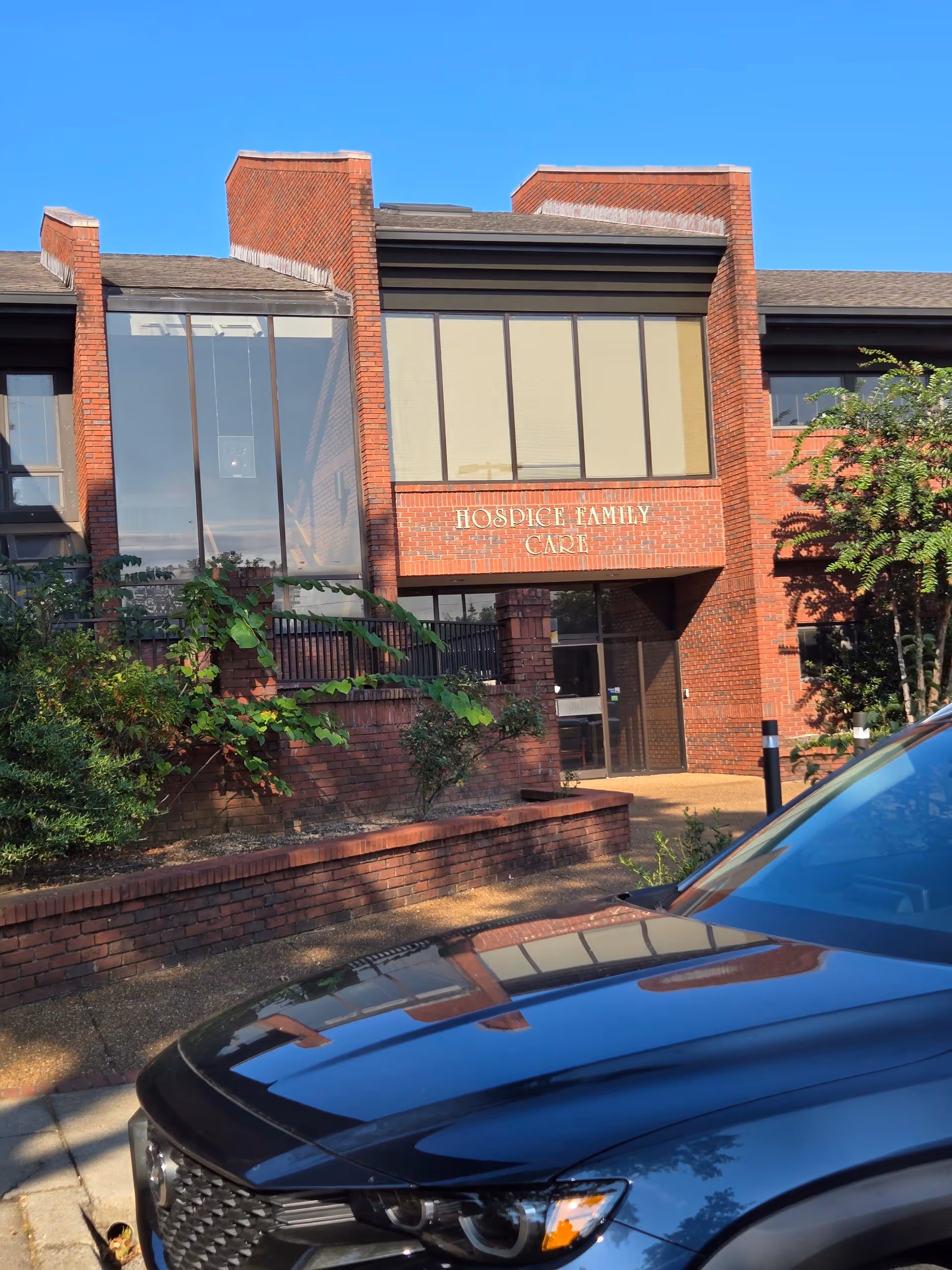 Brick building with large glass windows and a sign reading Hospice Family Care, entrance visible.