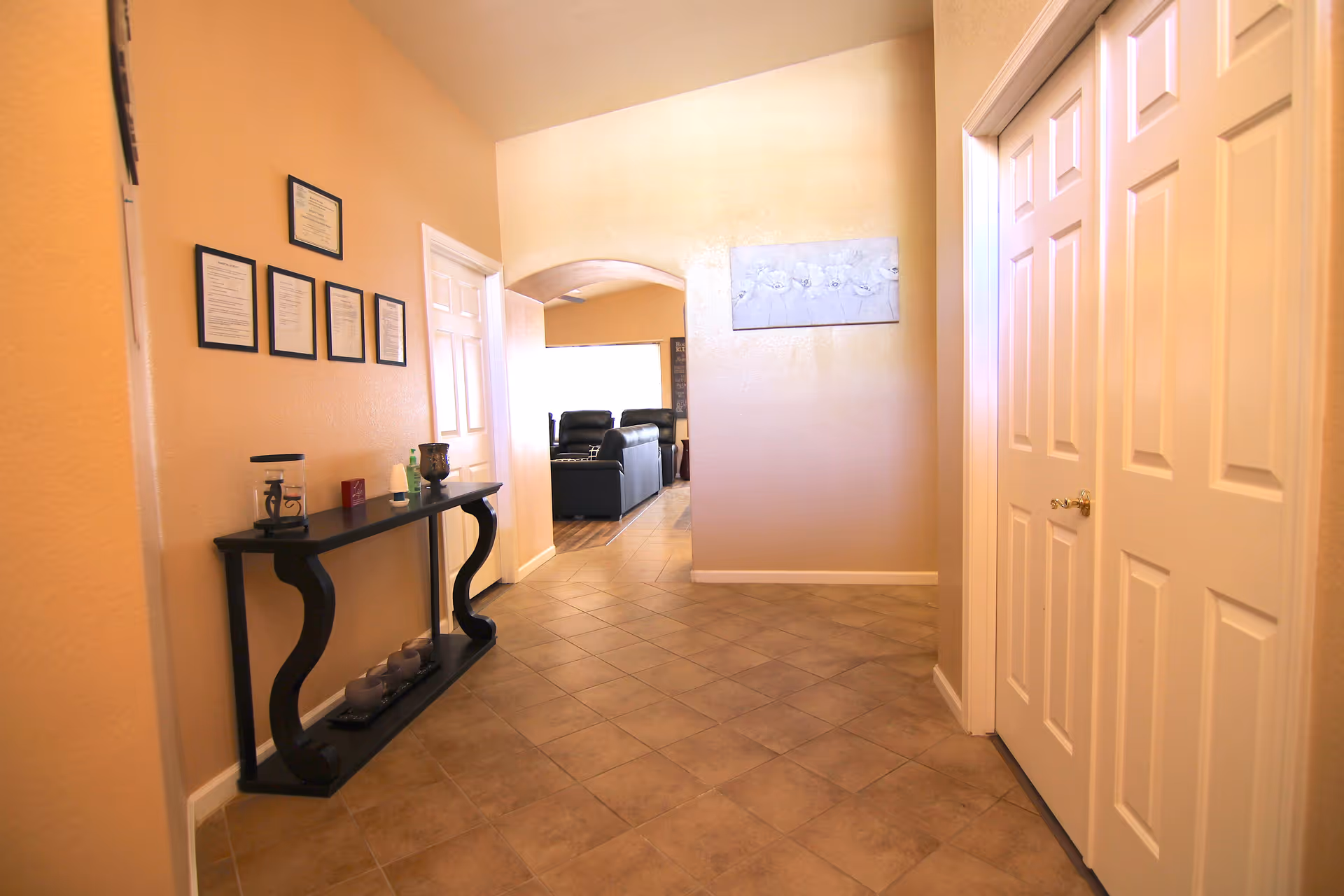 Interior hallway of Soft Touch Assisted Living with beige walls and tiled floor. On the left wall, there are framed certificates above a black console table holding decorative items. At the end of the hallway, an arched doorway leads to a living room area with black leather recliners. On the right side, there are white double doors and a wall with a floral painting.