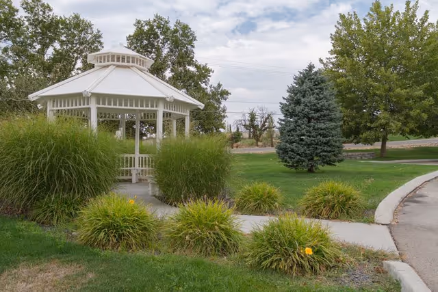 A white wooden gazebo surrounded by green bushes and trees in a grassy outdoor area with a curved concrete pathway leading to it under a partly cloudy sky.