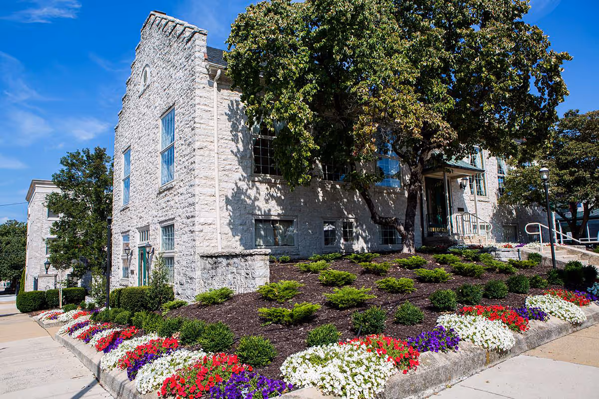 Stone building front with landscaped flowerbeds, trees, and a sidewalk leading to the entrance under a blue sky.