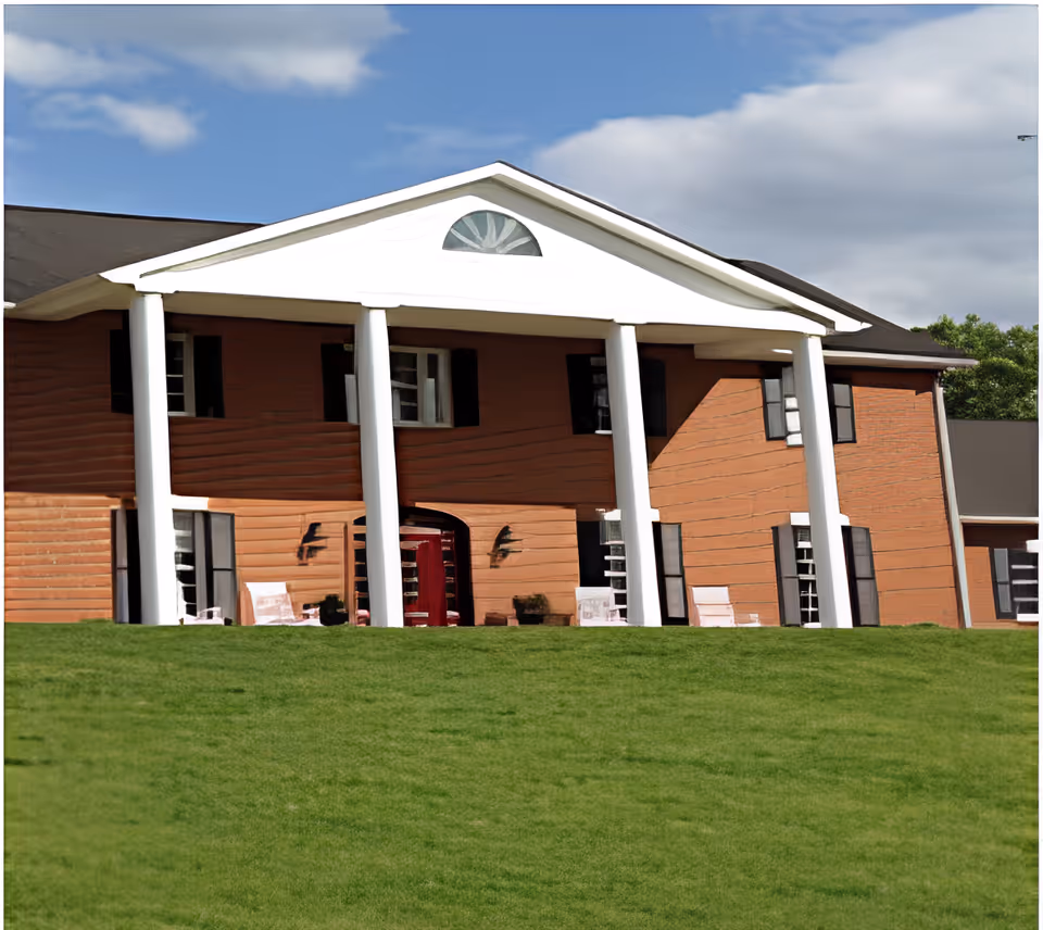 Two-story brick building with white columns and a large grassy lawn in front under a partly cloudy sky.