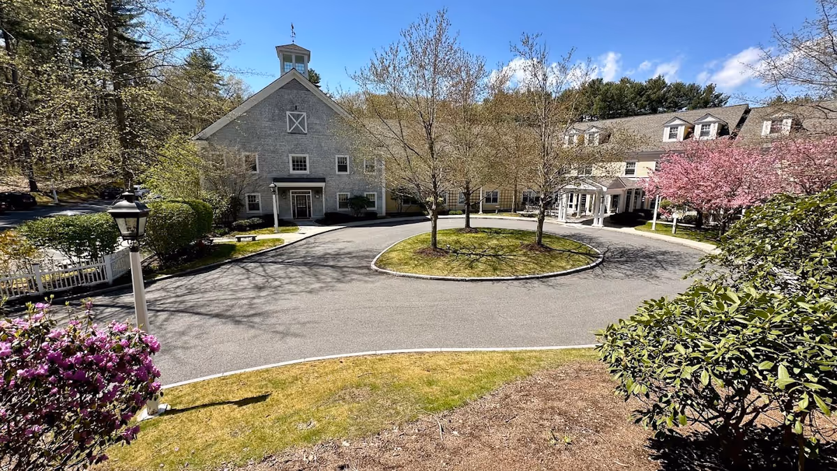 Front entrance of a senior living building with a circular driveway, landscaped trees and blooming shrubs under a blue sky.
