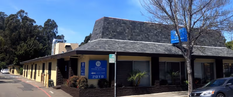 Exterior street view of the low-rise Bay Marina Post Acute building with blue signage, trees, and parked cars.