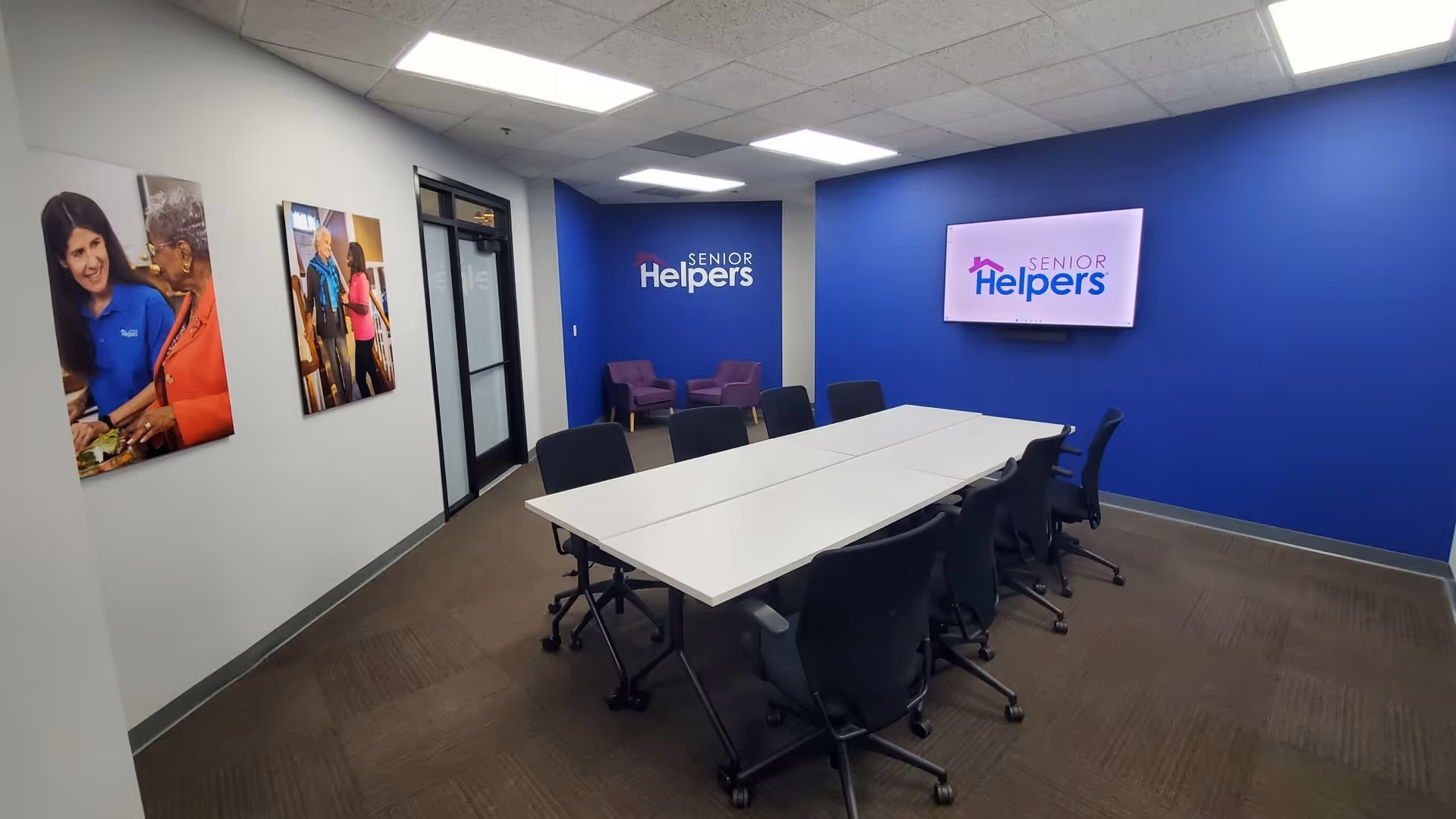 Interior conference room with a long white table, black chairs, and blue Senior Helpers wall.