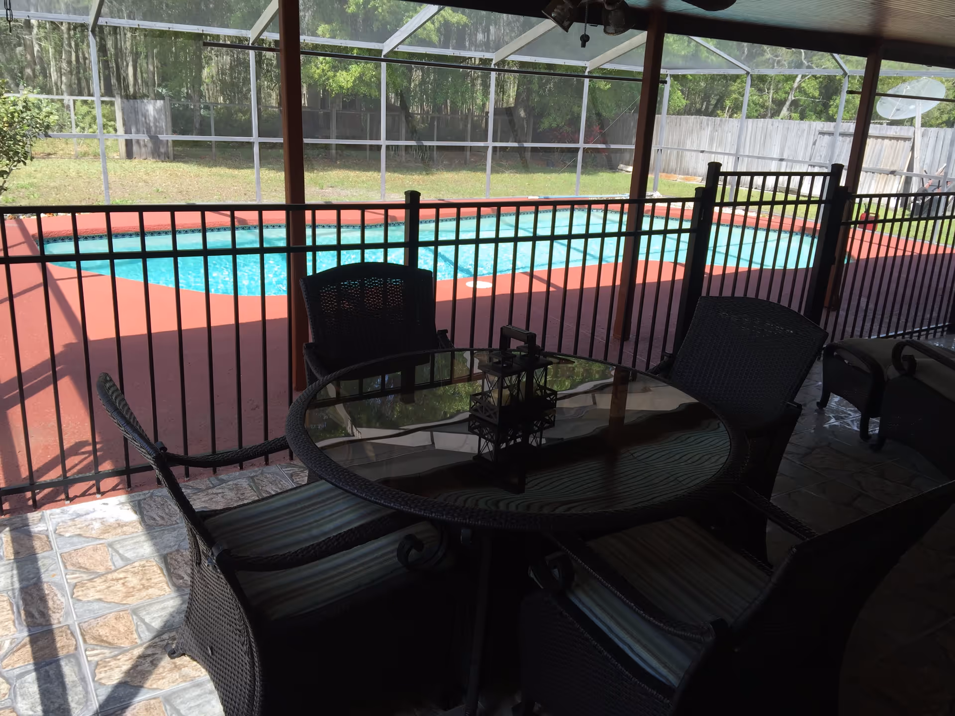 A covered outdoor patio area with a round glass-top table and four wicker chairs with cushions. Beyond the patio is a fenced swimming pool with clear blue water, surrounded by a red deck and enclosed by a screened structure. Trees and a wooden fence are visible in the background.