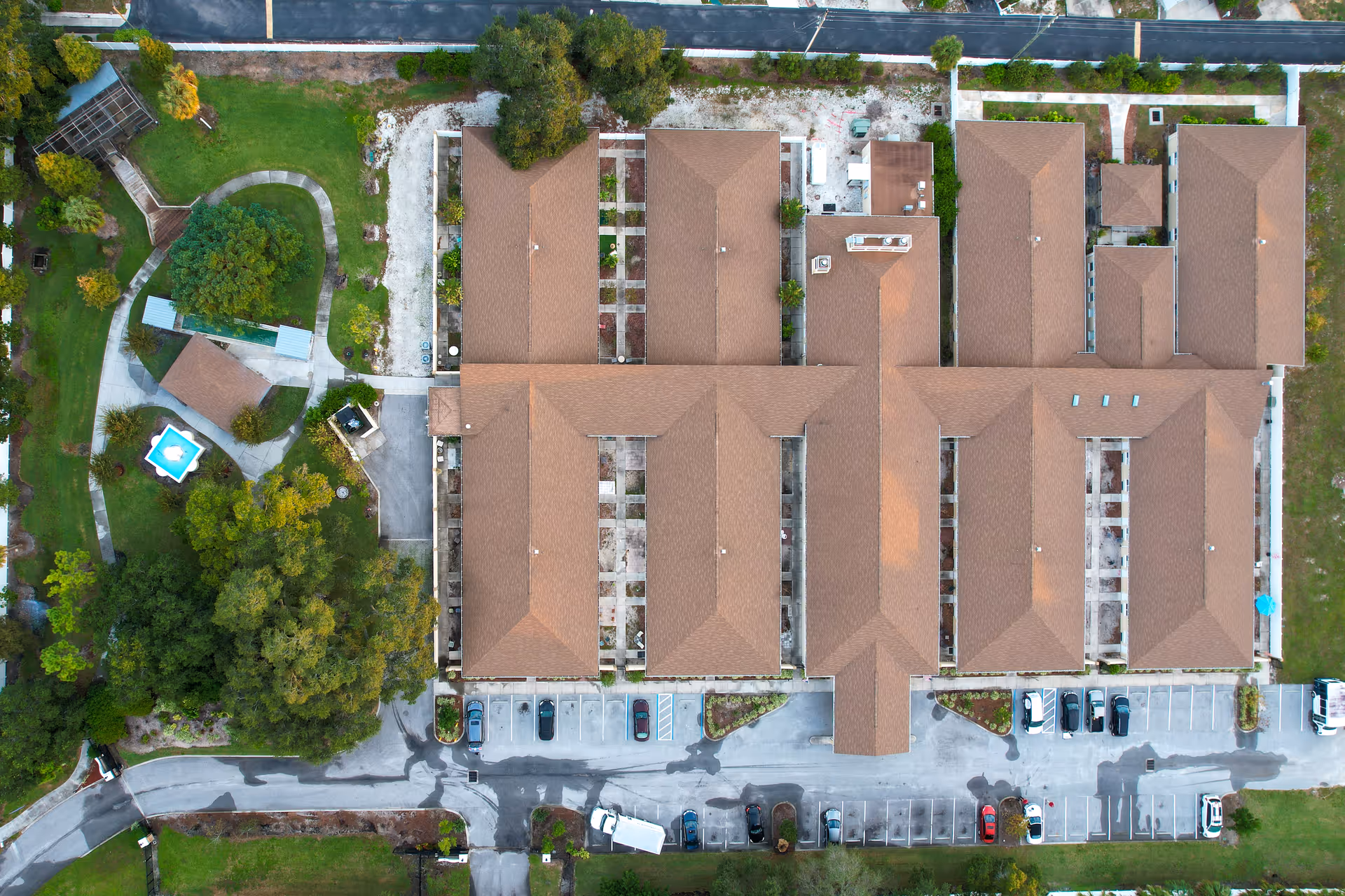 Aerial view of The Goldton at St. Petersburg senior living facility showing the building's roof, parking lot with cars, surrounding trees, pathways, and a small gazebo with a blue roof in a landscaped garden area.