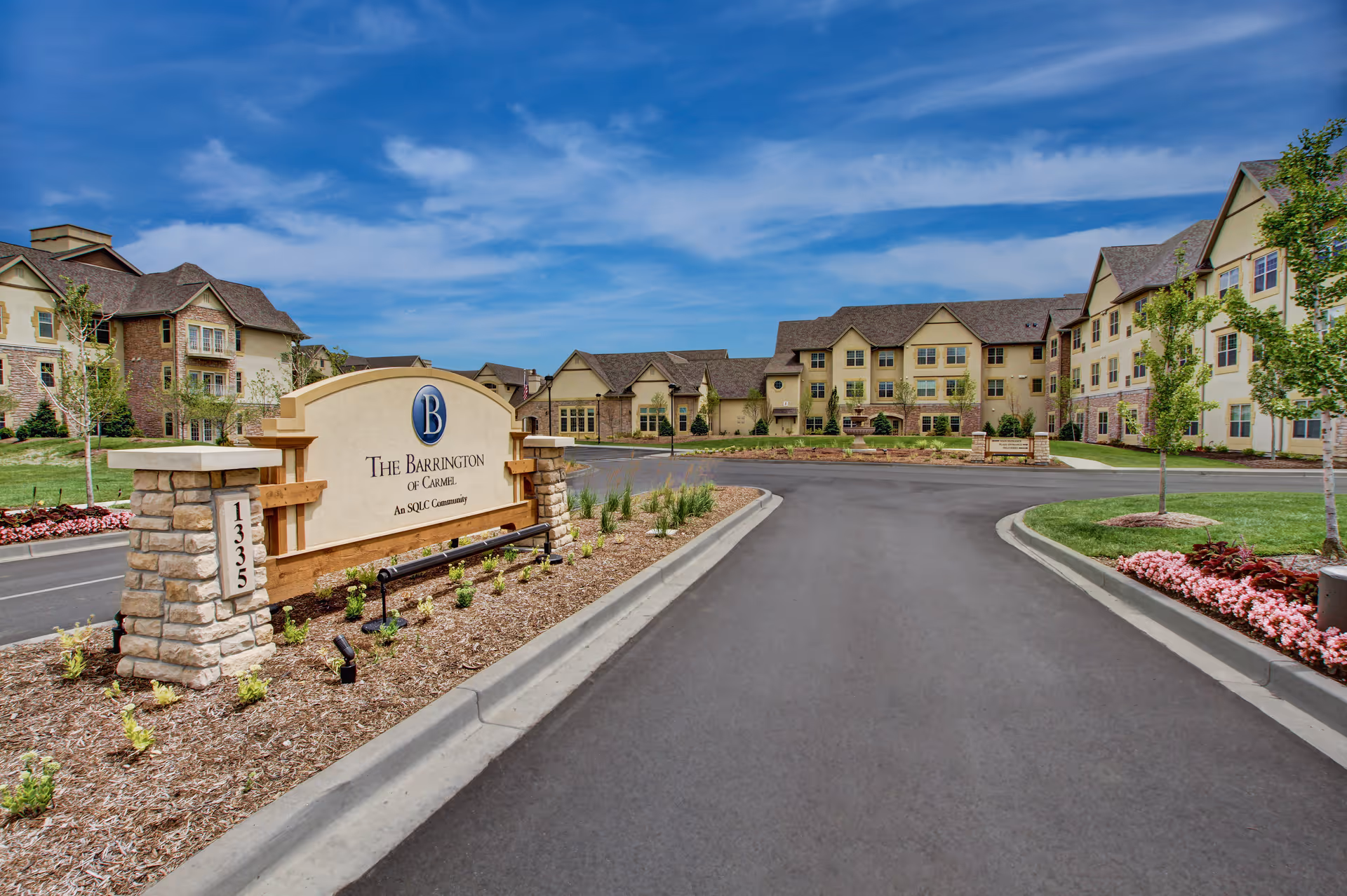Entrance to The Barrington of Carmel senior living community featuring a large stone and wood sign with landscaping, a paved driveway, and multi-story residential buildings under a partly cloudy blue sky.