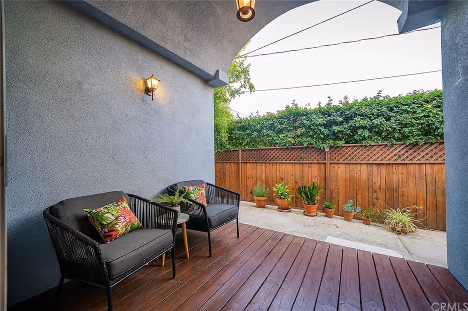 Covered outdoor patio area with two black wicker chairs with dark cushions and colorful floral pillows, a small wooden side table, a wall-mounted lantern light, wooden deck flooring, and a wooden fence with potted plants along the concrete ground outside.