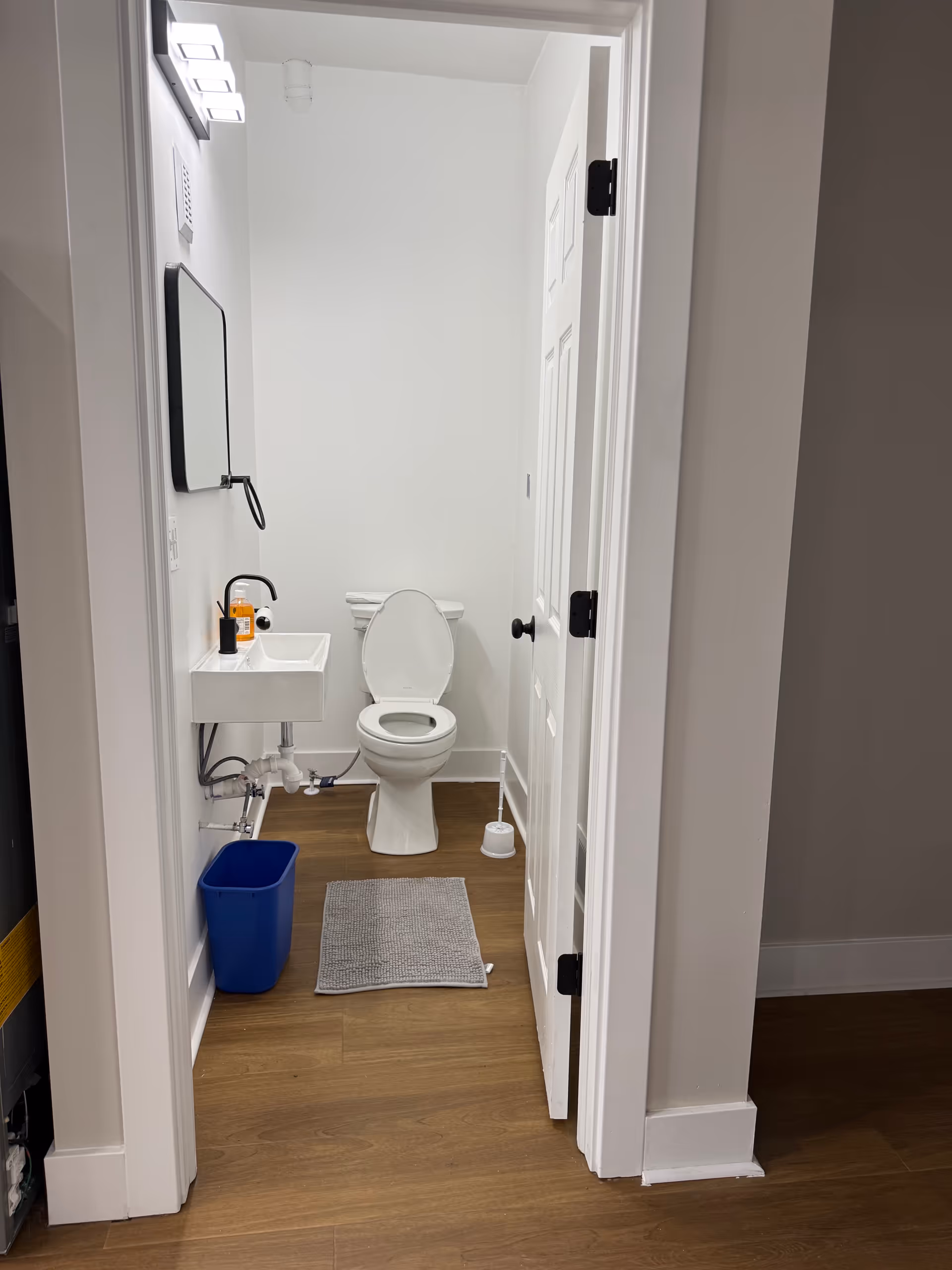 Compact white bathroom with toilet, pedestal sink, mirror, and a blue trash bin