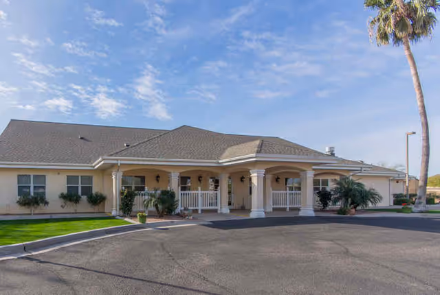 Front exterior of a single-story senior living building with a covered columned entrance, driveway, and a palm tree.