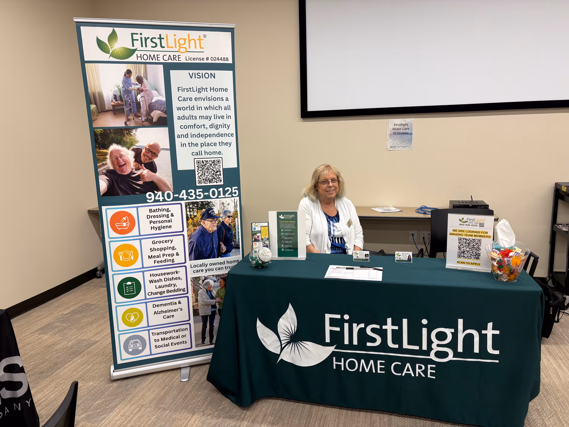 Indoor information booth for FirstLight Home Care with a smiling woman behind a green table.