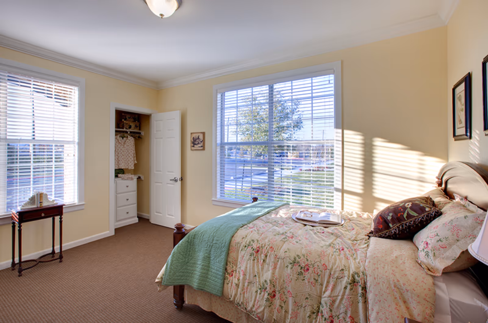 Sunlit bedroom with a floral-covered bed, large windows with blinds, a small side table, and an open closet.