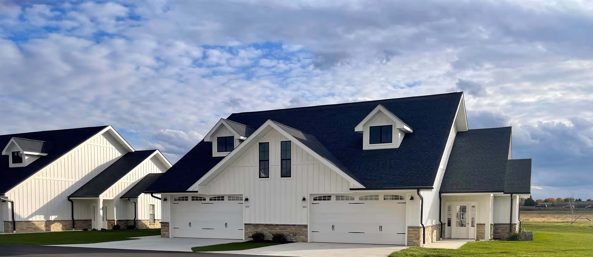 Exterior view of a modern senior living facility building with white siding, black roof, and attached garages under a partly cloudy sky. The building is surrounded by green grass and open space.
