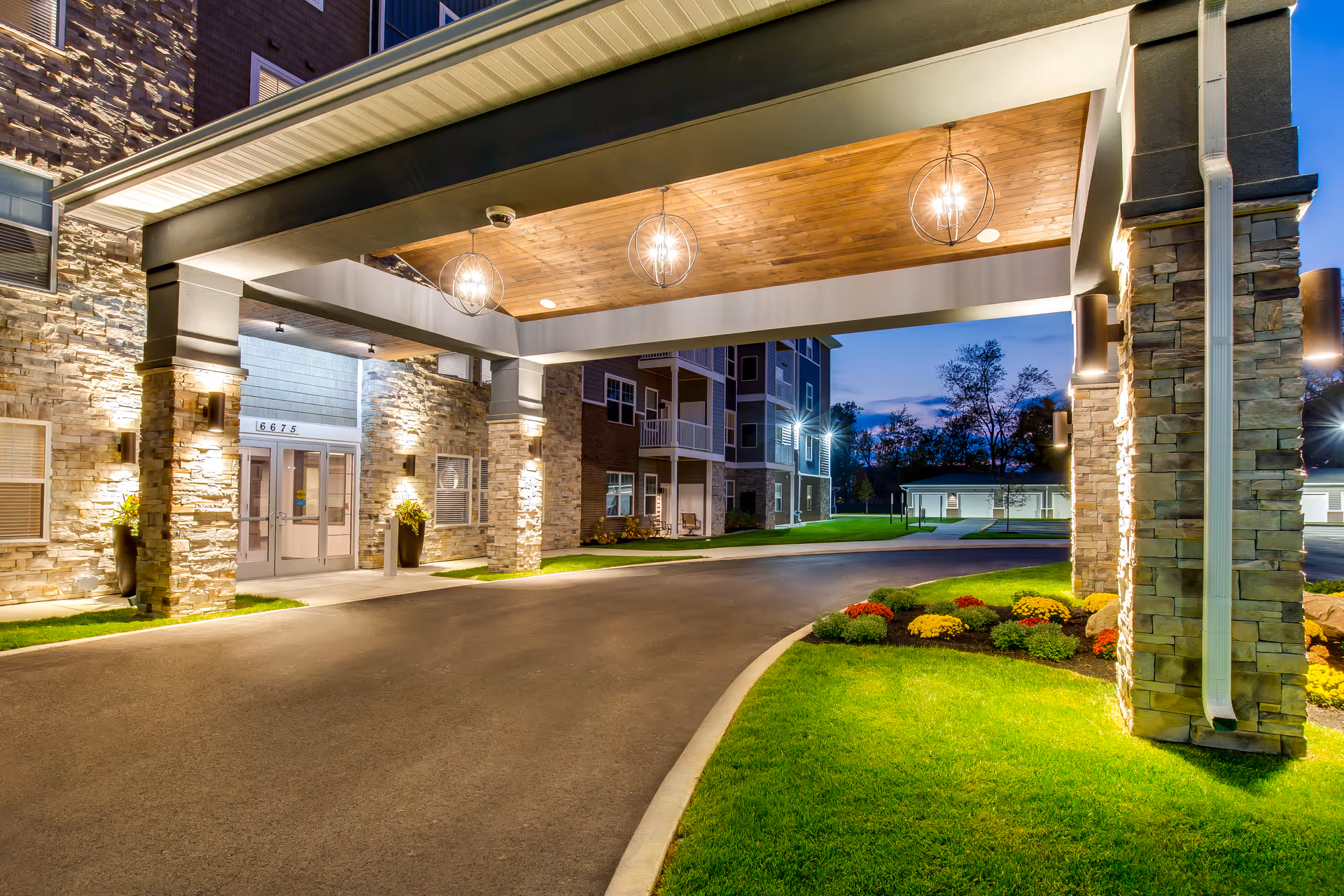 Covered entrance driveway and illuminated stone-faced facade of a senior apartment building with landscaping at dusk.