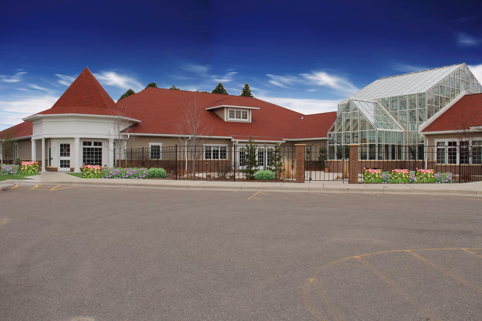 Exterior view of a senior living facility named Cottage On Forest by Three Links, featuring a building with a red roof, beige walls, a glass greenhouse structure, and a fenced garden area with flowers and shrubs under a blue sky.