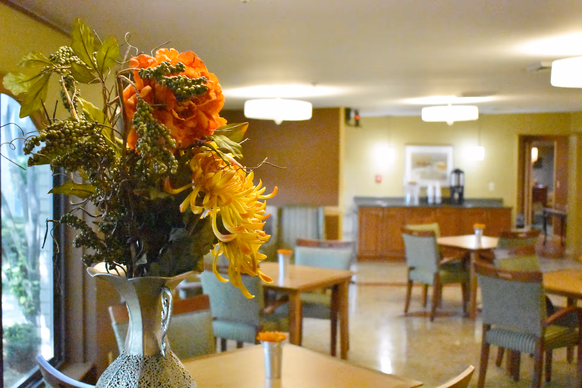 A senior living community dining area with wooden tables and cushioned chairs. A decorative vase with orange and yellow flowers is prominently placed on a table in the foreground. The room is softly lit with ceiling lights and has a warm, inviting atmosphere.