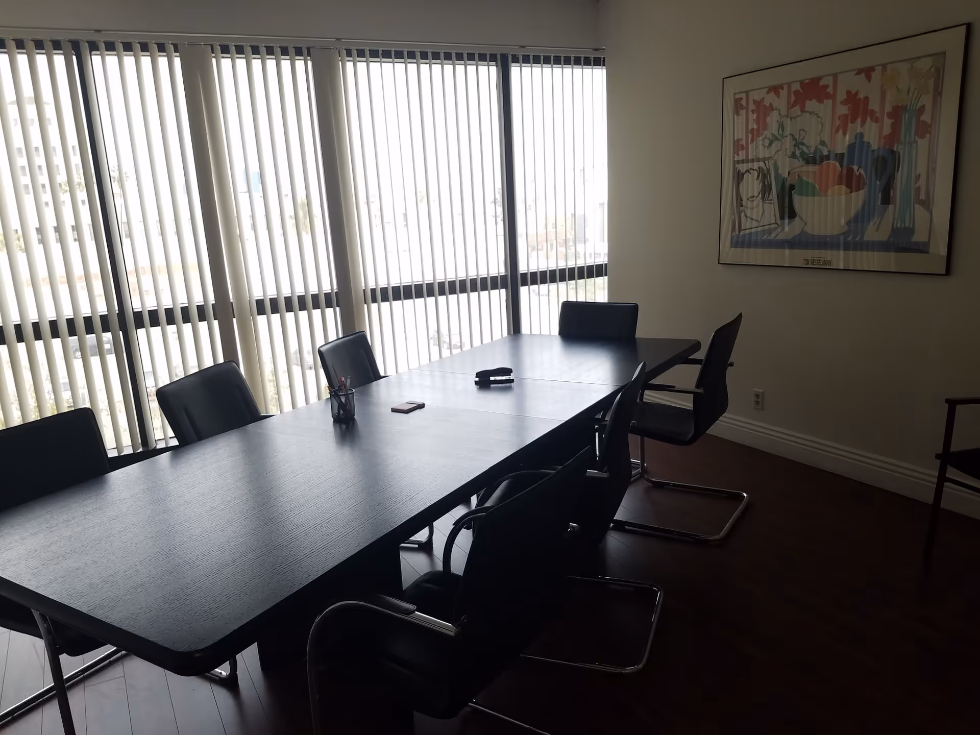 A modern interior conference room with a long table, black chairs, blinds, and wall art.