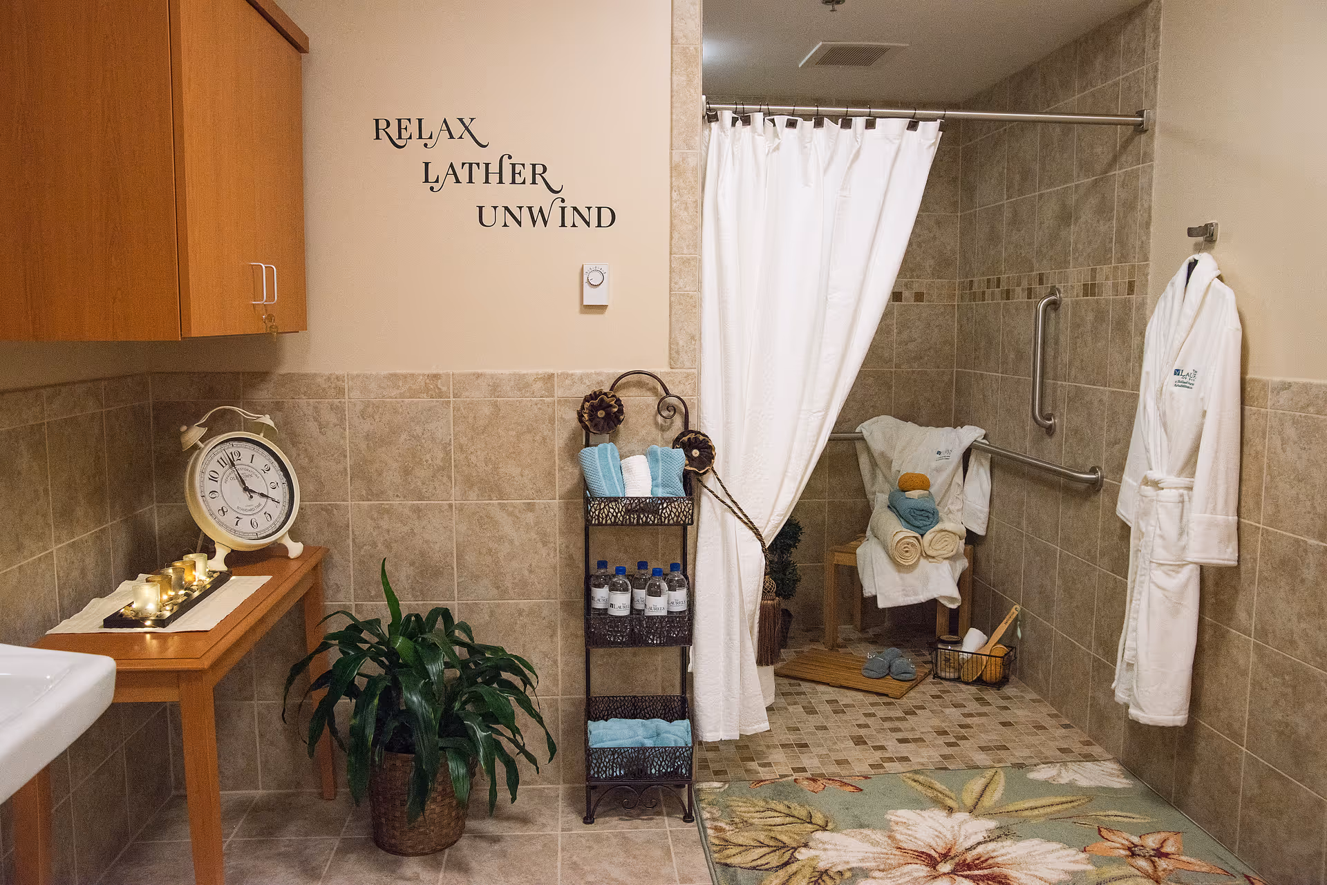 A bathroom with beige tiled walls and floor, featuring a walk-in shower with a white curtain. Inside the shower area, there are grab bars, a wooden bench with folded towels and bath items, and a basket with slippers. Outside the shower, there is a small table with candles and a clock, a potted plant, a metal rack holding rolled towels and water bottles, and a white bathrobe hanging on the wall. The wall has decorative text that reads 'RELAX LATHER UNWIND'.