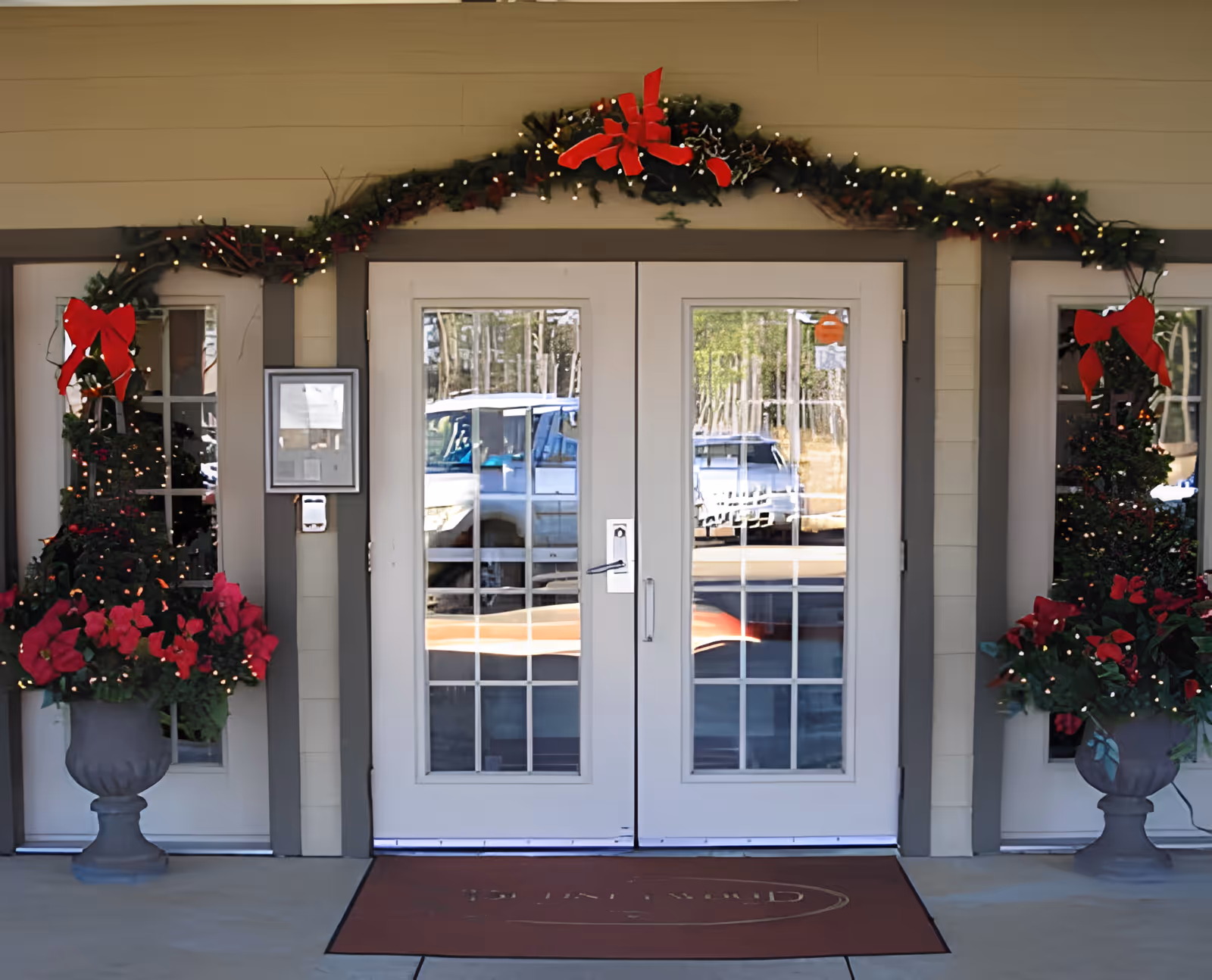 Entrance of Dudneywood Assisted Living decorated with Christmas garlands and red bows. Two white double doors with glass panes are centered, flanked by windows and large planters with red flowers and greenery. A red doormat is placed in front of the doors.