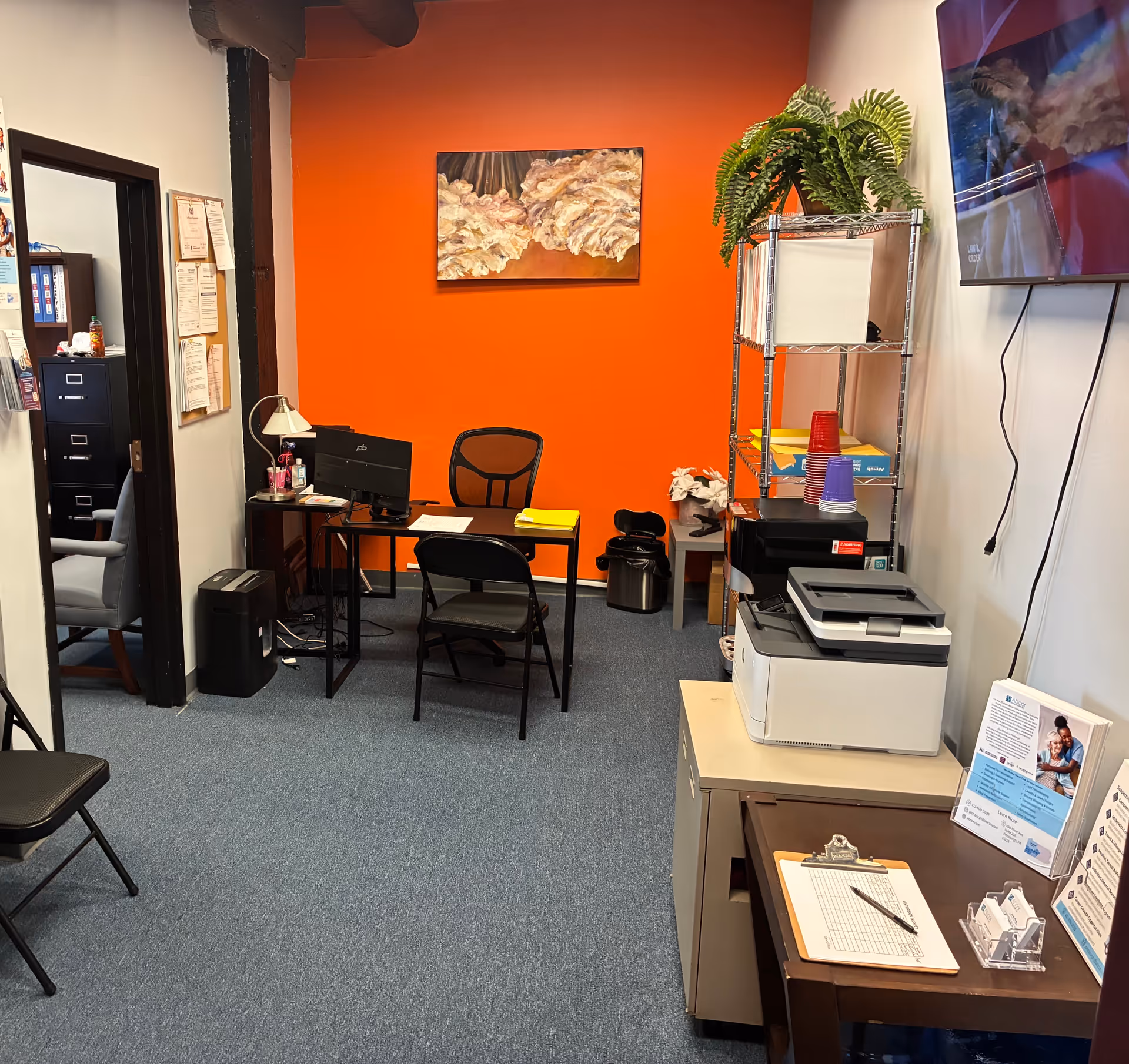 Office area with orange accent wall, desk, computer, printer, filing shelves, and plant.