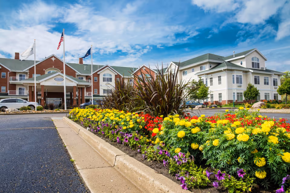 Exterior view of a senior living facility with a landscaped flower bed in the foreground featuring yellow, red, and purple flowers. The building has a combination of brick and light-colored siding with multiple windows and a covered entrance. Three flagpoles with flags are visible near the entrance under a partly cloudy blue sky.