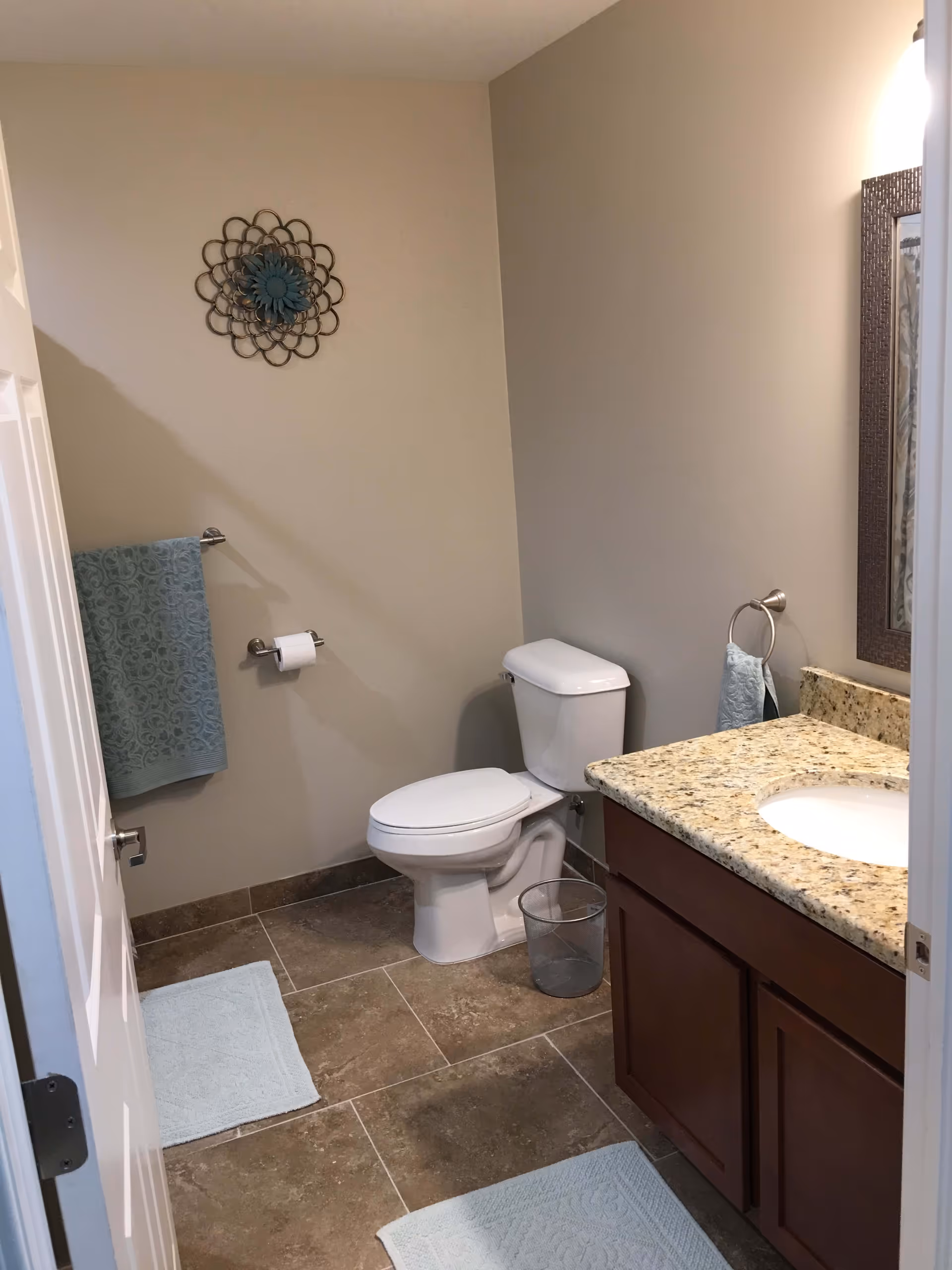 A clean bathroom with a white toilet, granite countertop with a sink, wooden cabinets, a large mirror, and beige tiled floor. There are light blue towels hanging on a towel rack and ring, a small trash bin next to the toilet, and a decorative metal wall art above the toilet paper holder.