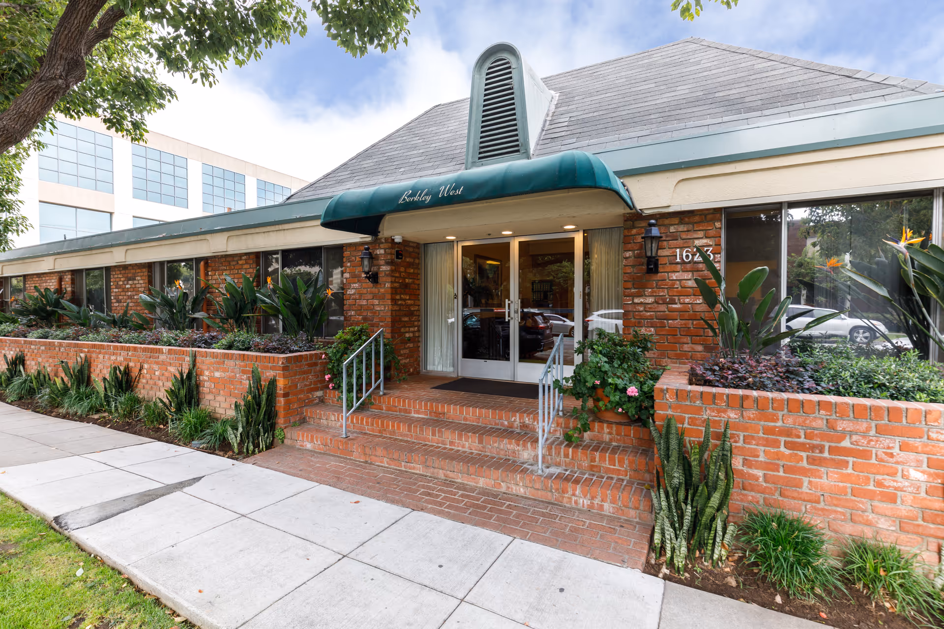 Exterior view of Berkley West Healthcare Center showing a brick building with a green awning above the entrance. The entrance has double glass doors with steps leading up to it, flanked by plants and greenery. The building number 1623 is visible on the right side of the entrance.
