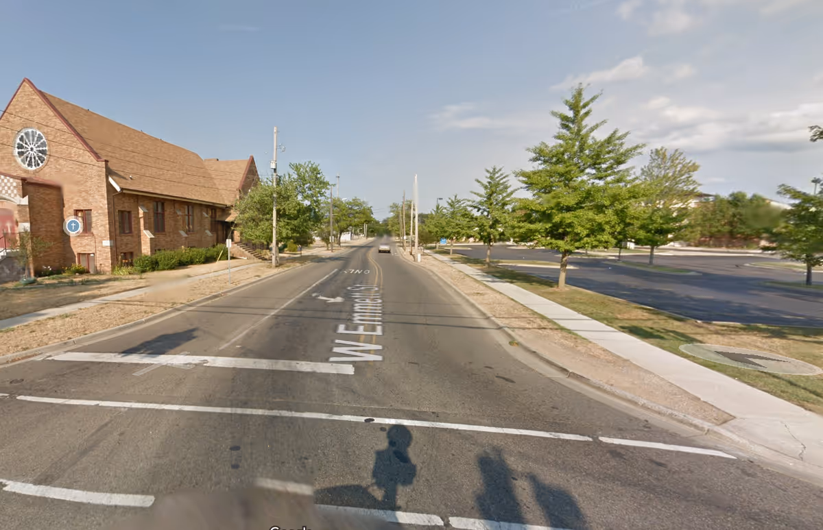 Street view of a brick building exterior with trees and a parking lot to the right.