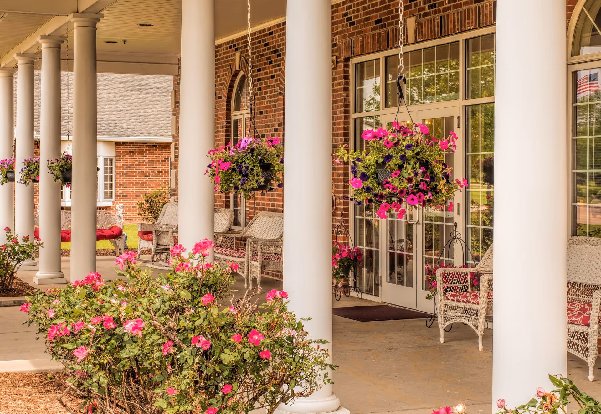 Covered front porch with white columns, hanging flower baskets, wicker chairs and glass entrance doors framed by brick.