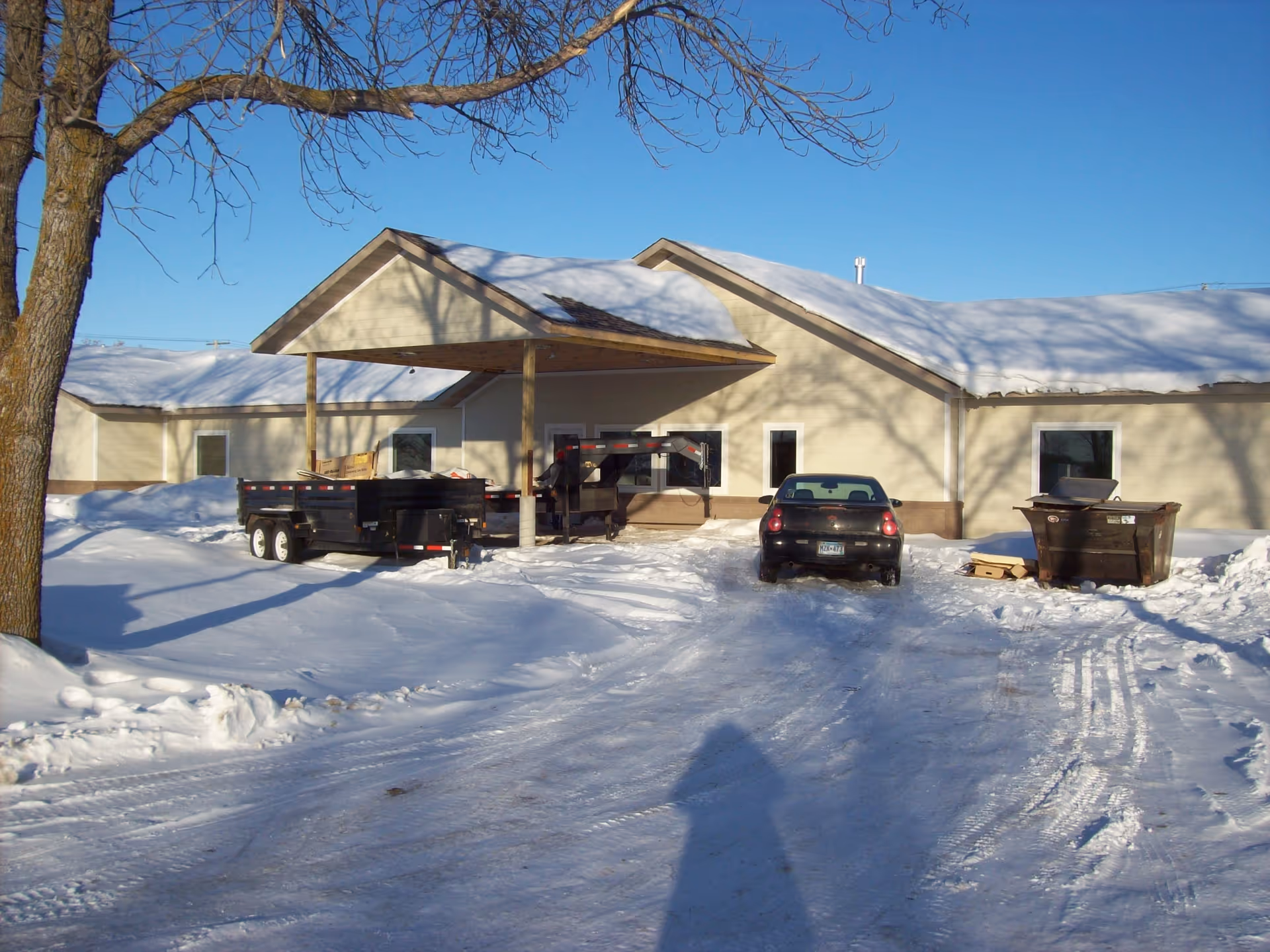 Snow-covered senior living center exterior with a driveway, car, and dump trailer.