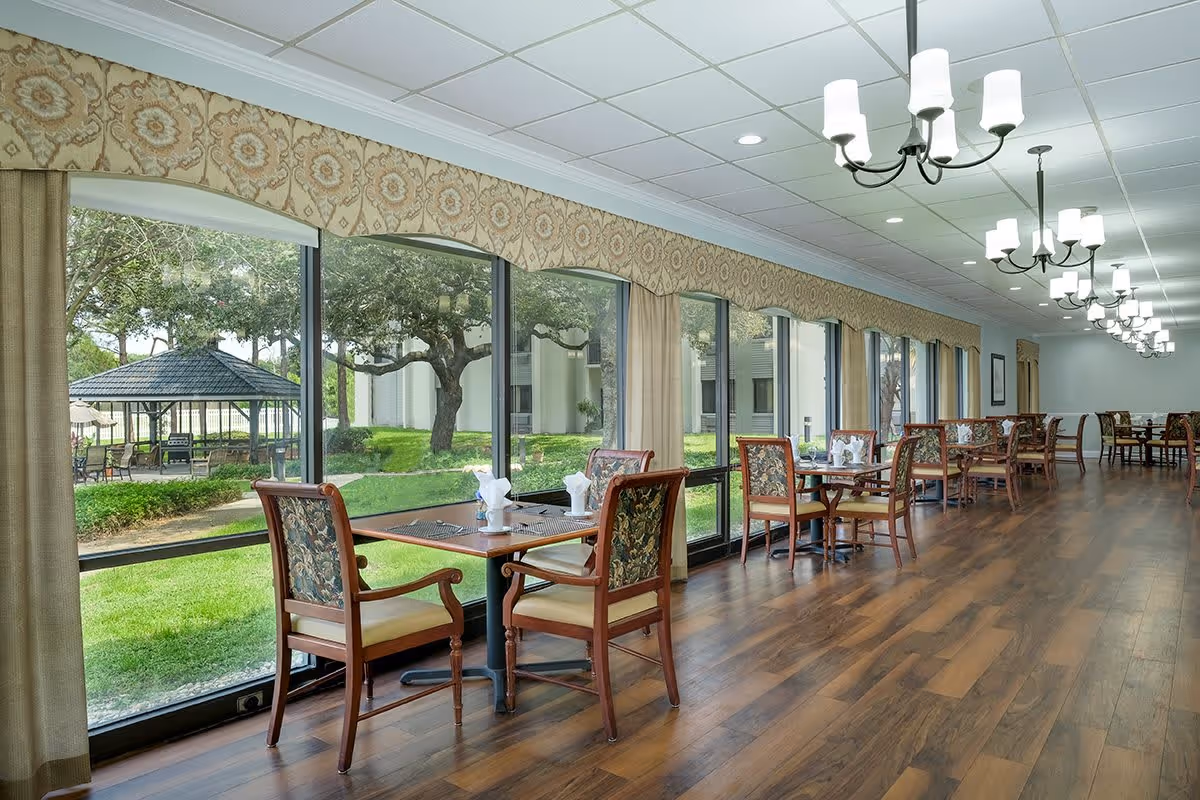 A long dining room with wooden floors and multiple tables set with napkins and placemats. The room has large windows with beige curtains and patterned valances, offering a view of a green outdoor area with trees and a gazebo. Several chandeliers hang from the ceiling providing lighting.