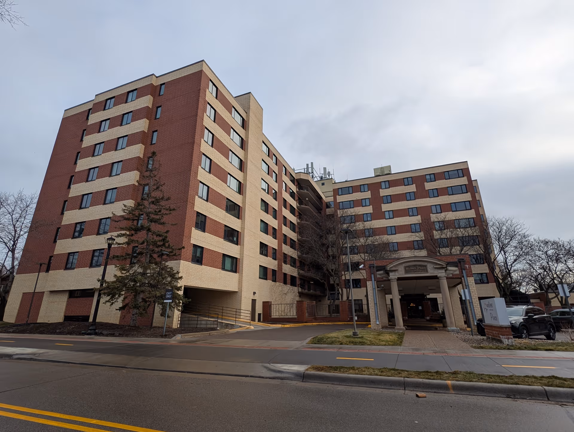 Exterior view of a multi-story senior living facility building named Walker Place by Vivie, with a covered entrance and several parked cars nearby under a cloudy sky.
