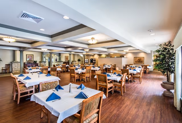 Spacious dining room with multiple tables set with white linens and blue napkins, wooden chairs, and warm lighting.
