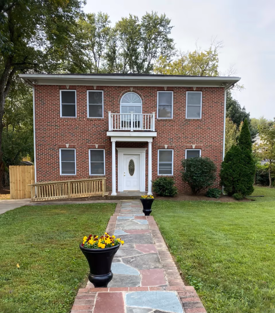 Two-story red brick building with a central white entrance, small balcony, and a flagstone walkway flanked by flower pots through a grassy lawn.