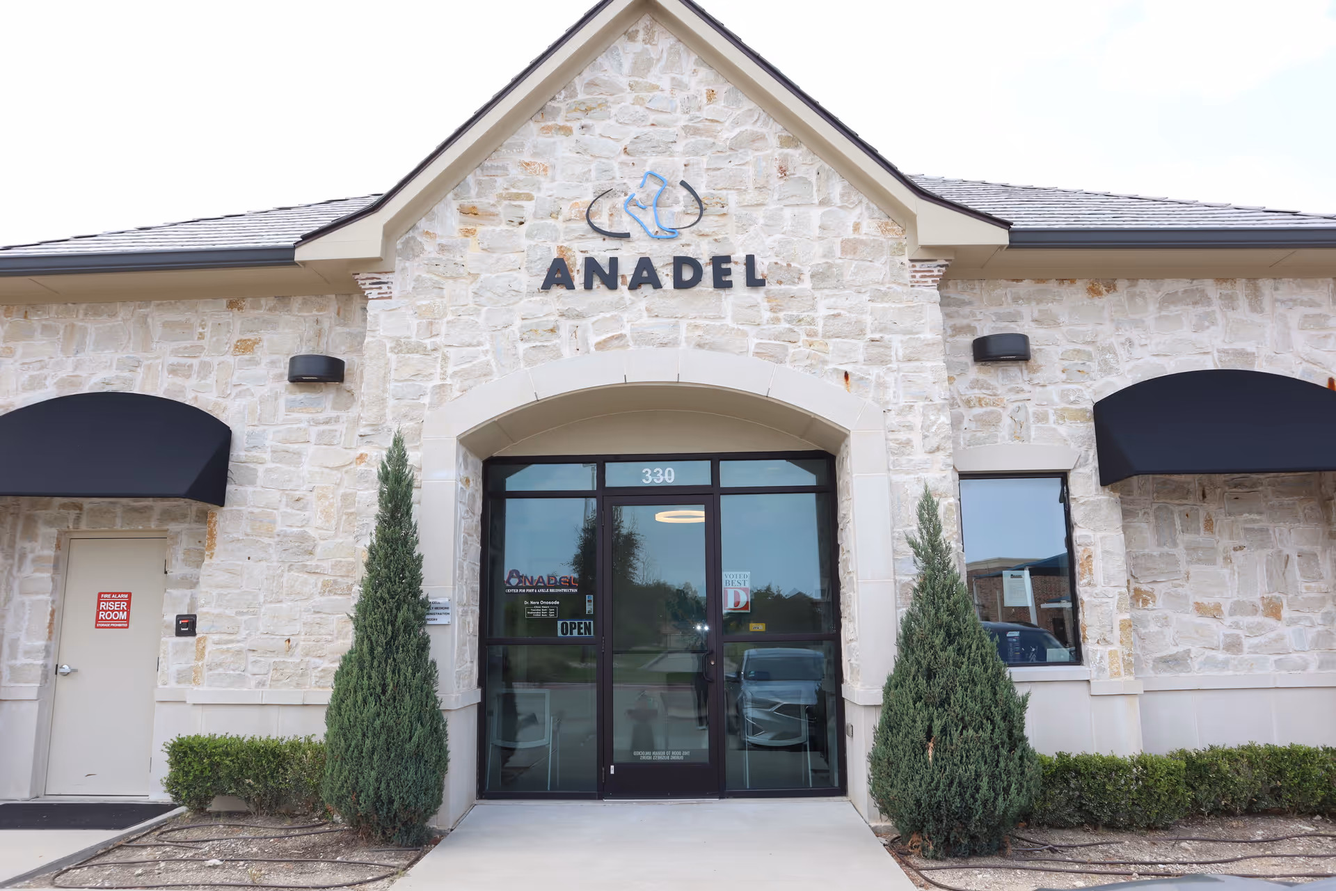 Stone-faced building exterior with glass doors and greenery flanking the entry.