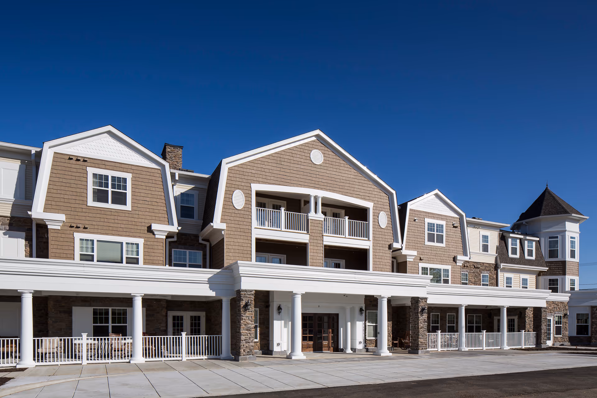 Exterior front view of a senior living facility building with multiple windows, balconies, and a covered entrance supported by white columns under a clear blue sky.