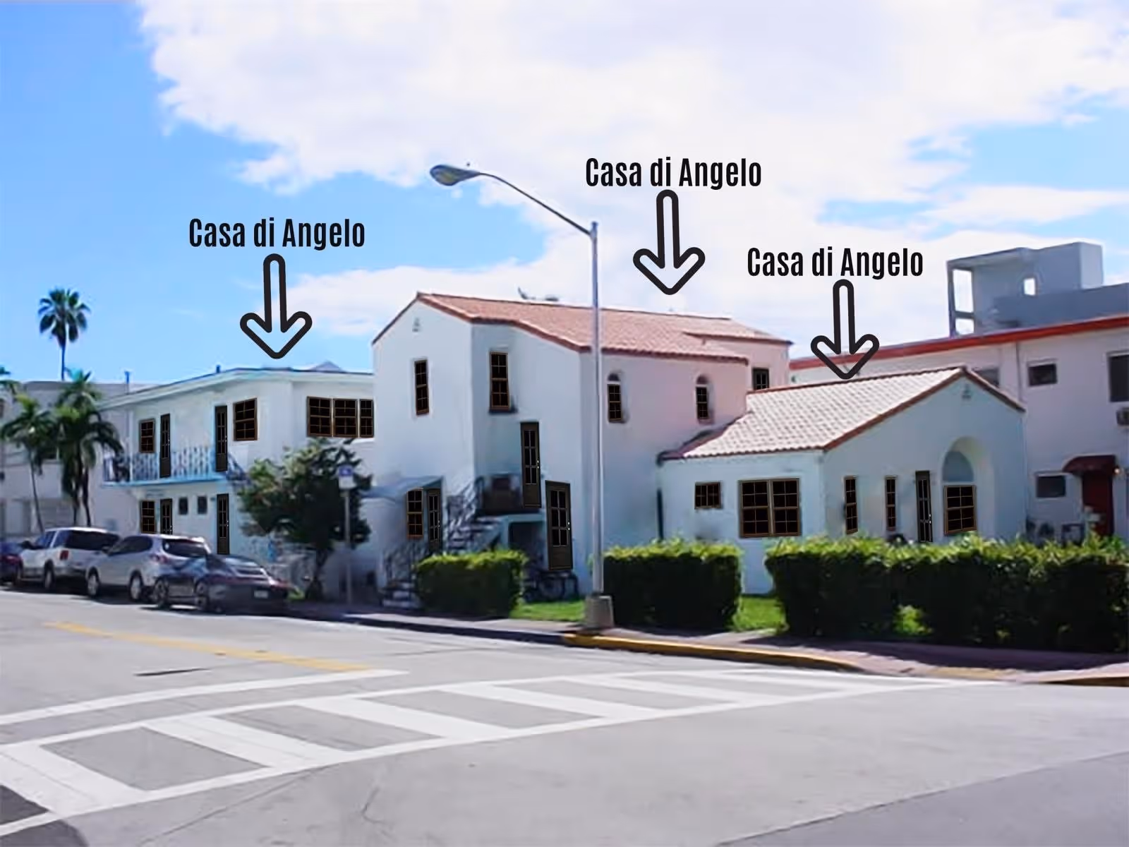 Exterior view of Casa di Angelo Friendship Suites showing multiple white buildings with red-tiled roofs along a street with parked cars and a streetlamp under a partly cloudy sky.