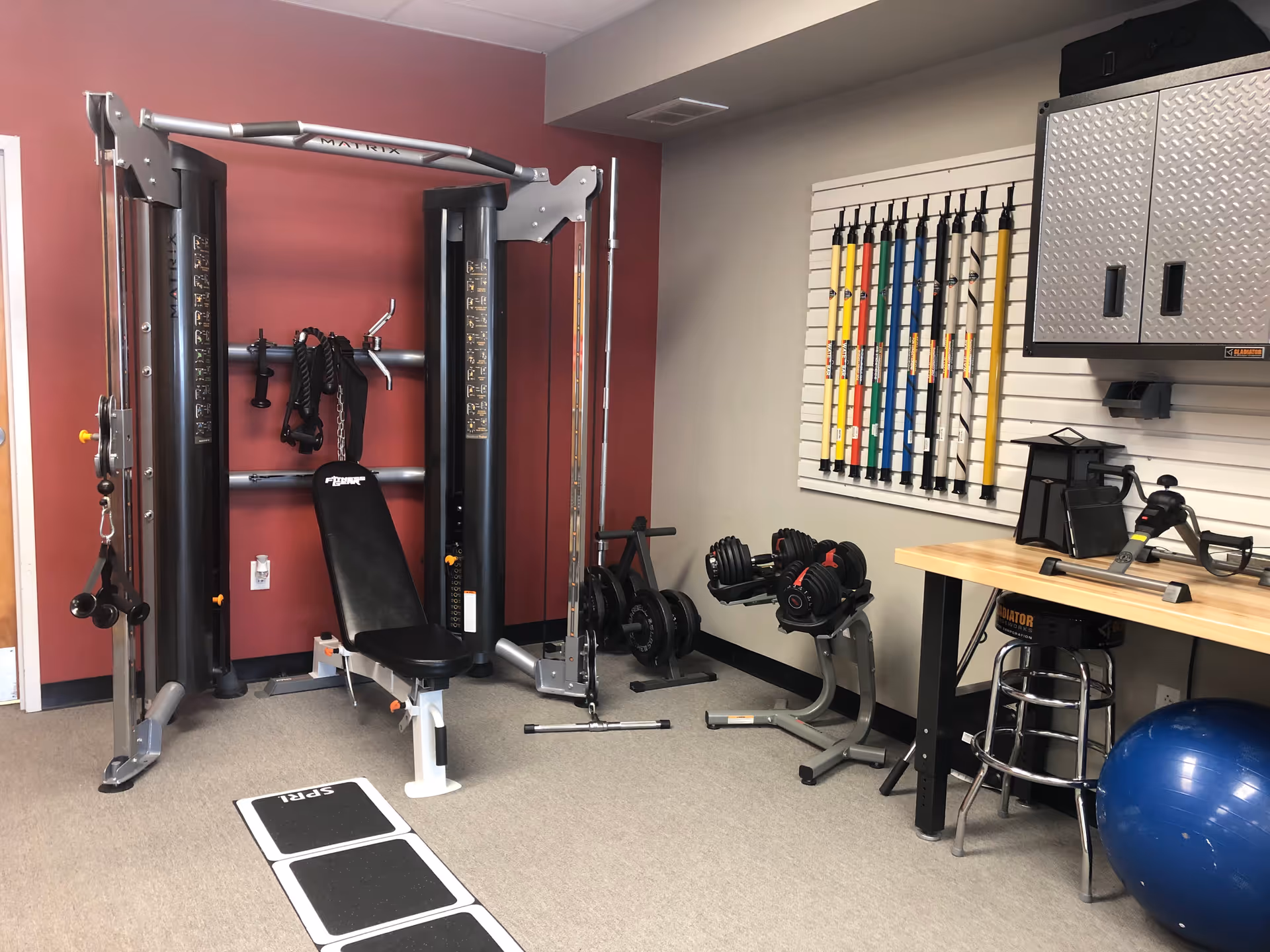 Small gym room with weight machines, benches, free weights, and a workbench in a senior facility.