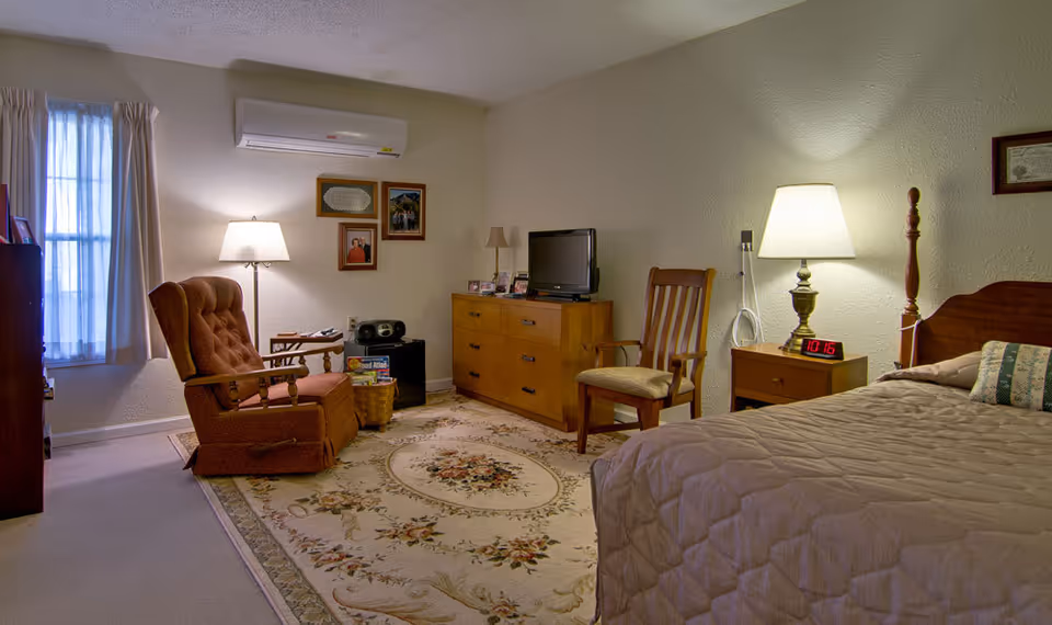 A cozy senior living bedroom with a bed covered in a beige quilt, a wooden nightstand with a lamp and digital clock, a wooden dresser with a TV on top, a wooden chair, and a cushioned armchair near a window with curtains. The room has a floral area rug and framed pictures on the wall.
