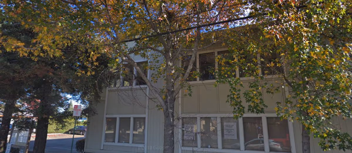 Exterior of a beige building with large windows partially shaded by trees.
