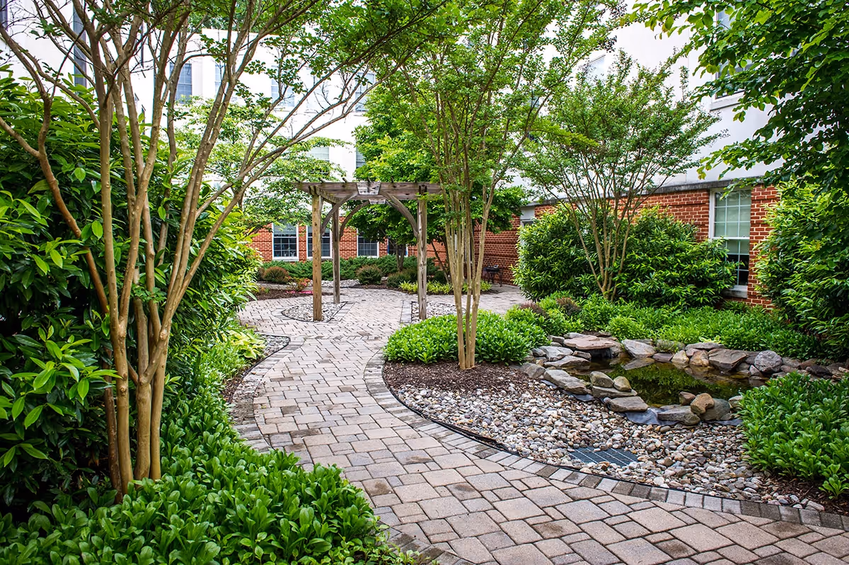 Curving brick-paved pathway through a landscaped courtyard with trees, shrubs, a pergola, and a small rock-lined pond beside a red brick building.