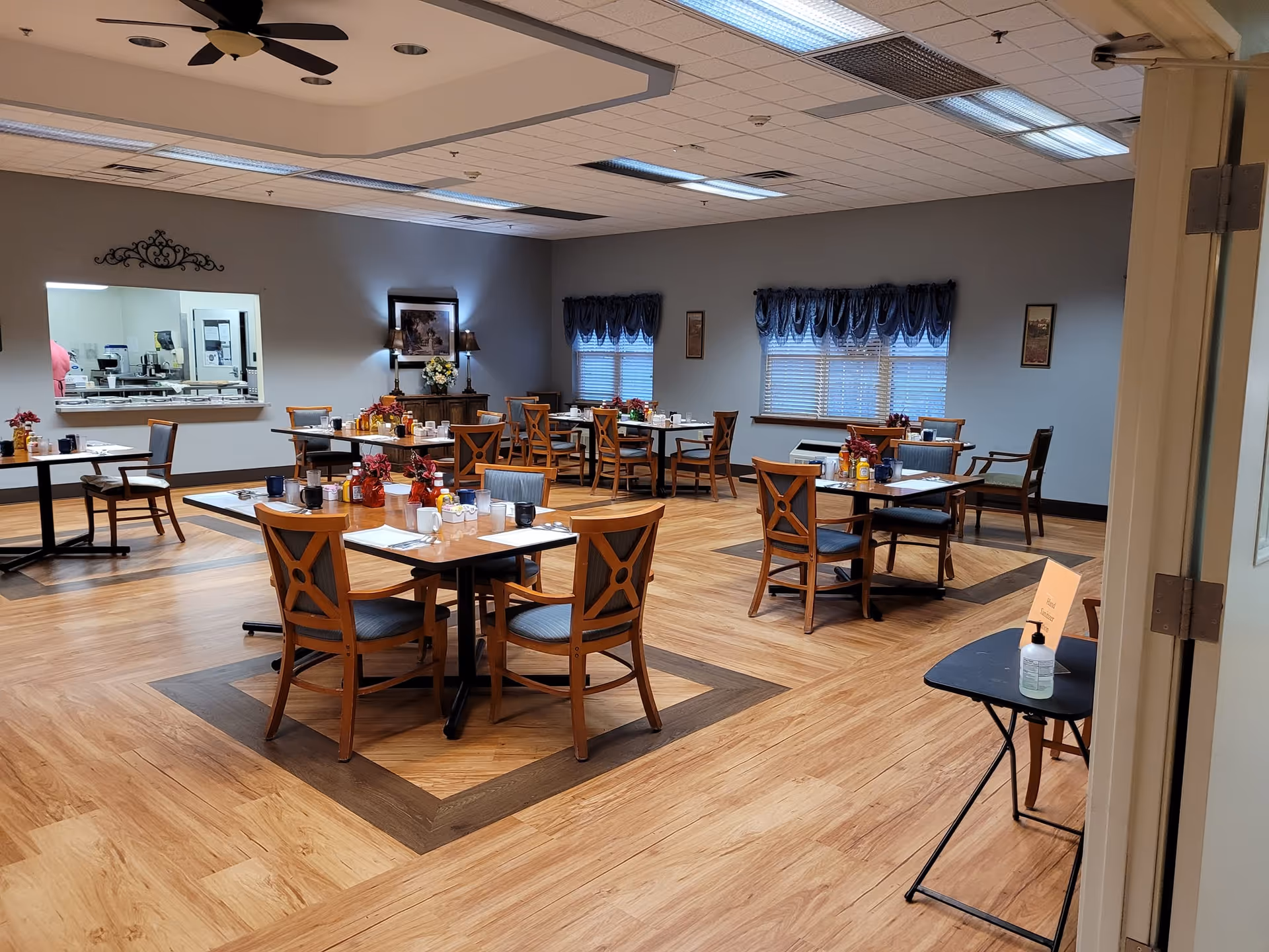 A spacious dining room with multiple wooden tables and chairs, and windows with blue curtains.