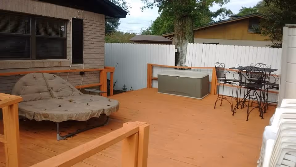 Wooden backyard deck with a cushioned round lounge chair, metal bistro table and chairs, a storage chest, and a white privacy fence.