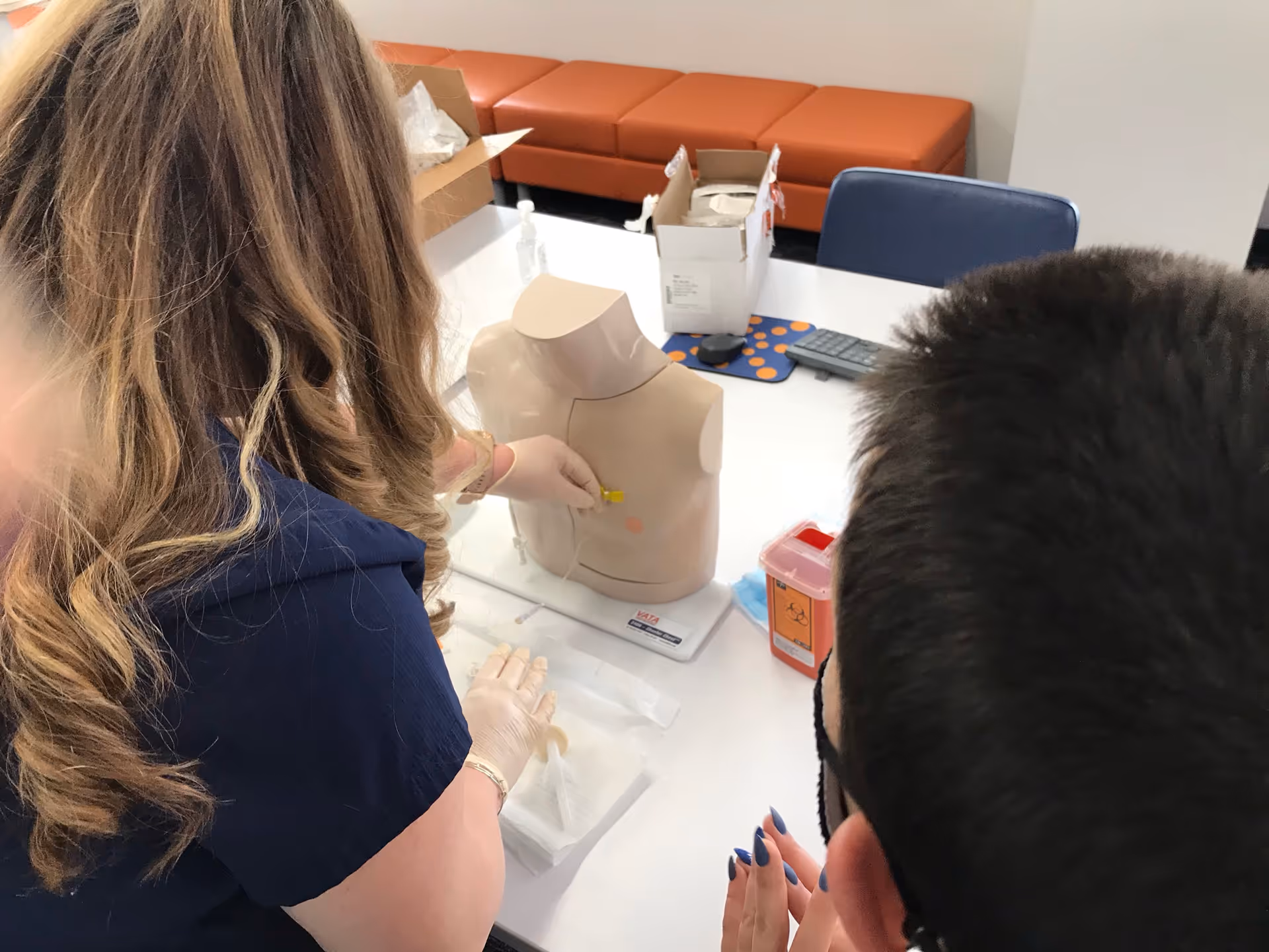 Two staff members practice medical training on a torso mannequin at a clinic table.
