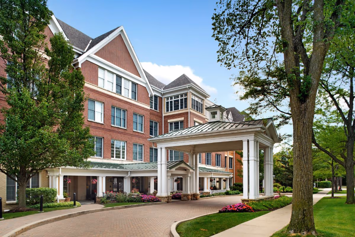 Front entrance of a multi-story brick senior living building with a covered porte-cochere, trees, and landscaped driveway.