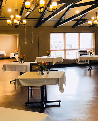 A dining room with several tables covered in white tablecloths, each adorned with small vases of colorful flowers. The room has wooden floors, large windows letting in natural light, and decorative ceiling beams with multiple chandeliers providing warm lighting.