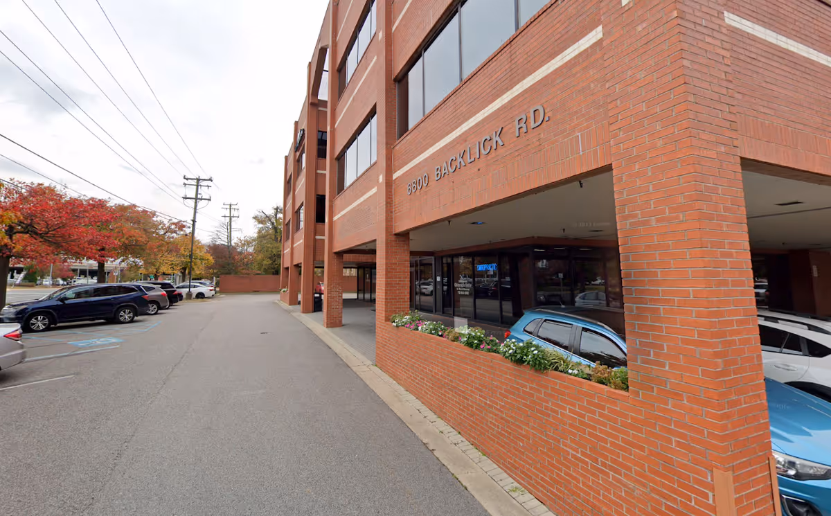 Brick building exterior with large windows, pillars, and parked cars along the street.