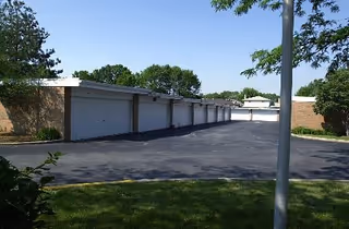 Exterior view of a row of closed white garage doors along a paved driveway with grass and trees under a clear blue sky.