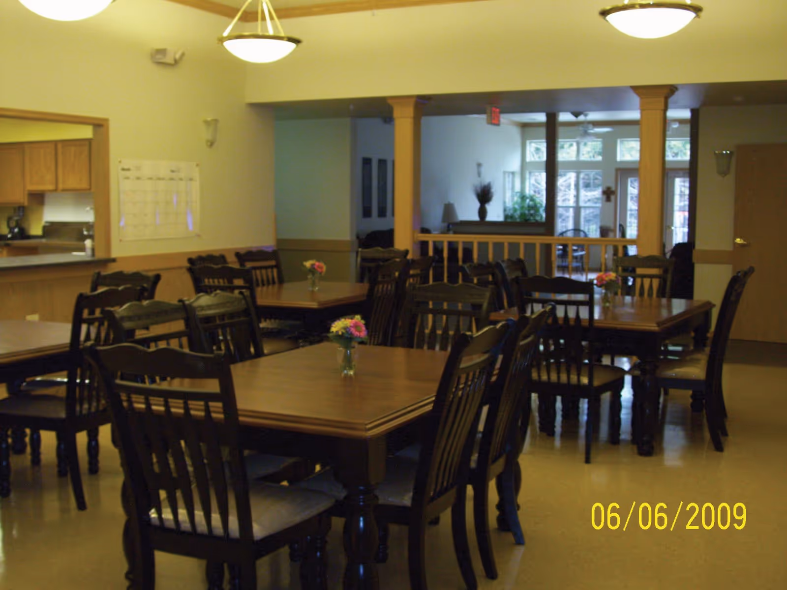 A dining room with several wooden tables and chairs arranged neatly. Each table has a small vase with flowers. The room has warm lighting with ceiling fixtures and a view into an adjacent kitchen area. There are large windows and a door in the background letting in natural light.